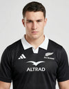 Maglia All Blacks Home 2025/26 Adidas primo piano busto frontale: colletto polo bianco con apertura V, badge ufficiale All Blacks con fern argentata in alto a destra, logo Adidas tre strisce bianco a sinistra, simbolo Altrad ad ali aperte al centro su fondo nero