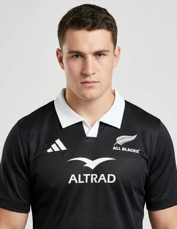 Maglia All Blacks Home 2025/26 Adidas primo piano busto frontale: colletto polo bianco con apertura V, badge ufficiale All Blacks con fern argentata in alto a destra, logo Adidas tre strisce bianco a sinistra, simbolo Altrad ad ali aperte al centro su fondo nero
