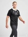 Maglia All Blacks Home 2025/26 Adidas vista tre quarti frontale sinistra: tessuto nero traforato, colletto polo bianco, logo Adidas bianco sul petto sinistro, stemma fern All Blacks a destra, sponsor Altrad al centro, indossata con pantaloni grigio antracite