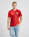 Maglia Canada Home 2026 Nike indossata da modello, vista frontale intera: rosso brillante con grafica geometrica a stella sul petto, stemma Canada Soccer a destra e swoosh Nike bianco a sinistra, colletto a V, polsini con banda nera, abbinata a jeans chiari